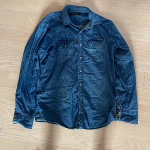 Blå jeansskjorta från Replay - Snygg blå jeansskjorta från Replay med klassisk krage, tryckknappar och två bröstfickor med lock. Skjortan har långa ärmar och en avslappnad passform. Perfekt för dig som gillar en cool och tidlös stil.