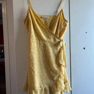 Gul blommig omlottklänning Hollister - Superfin gul kortklänning från Hollister med blommigt mönster, omlottmodell och volangkant nertill. Klänningen har smala axelband och knyt i sidan för en snygg passform. Perfekt för varma dagar och ger en fräsch sommarkänsla.