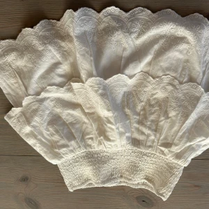 Vit broderad kortkjol från Kappahl - Superfin vit kortkjol från Kappahl med bred smock i midjan och broderade blommor längs den vågiga kanten. Kjolen är i bomull och har en romantisk, luftig känsla – perfekt för varma dagar.