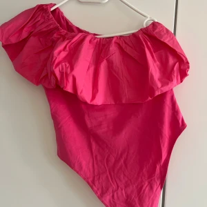 Rosa topp - Snygg rosa topp med stor volang upptill och asymmetrisk axel. Perfekt för dig som vill sticka ut på stranden eller vid poolen. Materialet känns lätt och bekvämt mot huden. Supersnygg och trendig design!