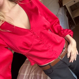 Röd blus med knappar från Kappahl - Snygg röd blus i glansigt material från kappahl med v-ringning och dekorativa knappar framtill. Blusen har långa puffärmar med resår vid ärmslut och en croppad passform. Perfekt för dig som vill sticka ut lite extra.