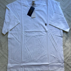 Vit t-shirt från Polo Ralph Lauren - Vit t-shirt från Polo Ralph Lauren med klassisk svart broderad logga på bröstet. T-shirten har rund hals, korta ärmar och är gjord i mjuk bomull. Slim fit-modell som passar perfekt till jeans eller shorts.