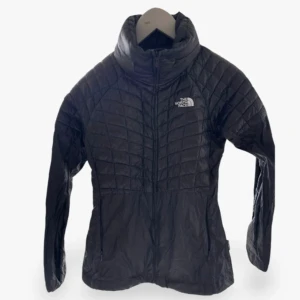 Svart quiltad dunjacka The North Face - Svart quiltad dunjacka från The North Face med hög krage och dragkedja framtill. Jackan har diskret logga på bröstet och är tillverkad i lätt, glansig polyester. Perfekt för kyliga dagar och enkel att bära med sig. Snygg passform och klassisk look.