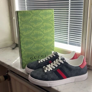 Gucci sneakers med röd detalj - Gucci sneakers i mörkblått med klassiskt GG-mönster, vita snören och röd samt marinblå rand på sidan. Baksidan har röd hälkappa med guldfärgad Gucci-logga. Snygg vit sula med Gucci-mönster undertill. Skorna levereras med originalkartong och dustbag. Tyvärr är boxen lite sönder! Skorna är använda 3-5 gånger 