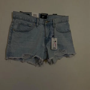 Ljusblå jeansshorts från 157 - Säljer ett par ljusblå jeansshorts från 157 med råa, fransiga kanter och slitna detaljer framtill. Klassisk femficksmodell med knapp och dragkedja. Perfekta för varma dagar och en avslappnad look.