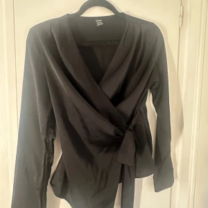 Svart omlottopp med knyt från  - Snygg svart omlottopp från  med långa ärmar och knyt i midjan. Toppen har en djup v-ringning och är gjord i ett mjukt, lätt material som faller fint. Perfekt för dig som gillar stilrena och trendiga plagg.