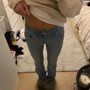 Ljusblå jeans med korsdetalj - Säljer ett par ljusblå jeans med snygga detaljer och bling på fickorna. Baksidan har en stor korsapplikation med stenar och broderier. Jeansen har låg midja och raka ben, perfekt för dig som gillar statement-plagg.