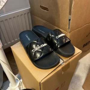 Svarta Calvin Klein tofflor - Svarta sandaler från Calvin Klein med quiltad rem och logga i vitt framtill. Snygg och enkel design med platt sula, perfekt för sommardagar eller att chilla hemma.