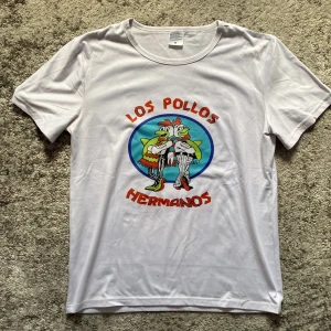 Los Pollos Hermanos vit t-shirt M - Vit t-shirt med färgglatt Los Pollos Hermanos-tryck på bröstet. Motiv med två tecknade kycklingar i cirkel, röd text och detaljer i blått, grönt och orange. Rund hals och korta ärmar. Tillverkad i mjuk bomull, perfekt för dig som gillar popkultur och unika prints.