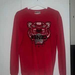 Röd sweatshirt med tiger från Kenzo - Säljer en röd sweatshirt från Kenzo med ikonisk broderad tiger på bröstet och texten 'Kenzo Paris'. Tröjan har rund hals, långa ärmar och är tillverkad i mjuk bomull. Perfekt statement-plagg för dig som gillar att sticka ut. Knappt använd men legat i garderoben ett tag, storleken på tröjan är Small och därför försöker jag bli av med den
