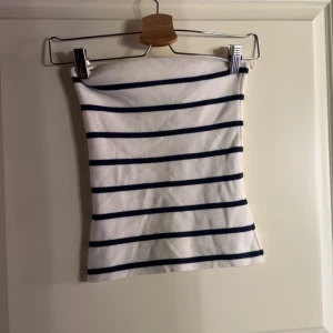 Randig vit och marinblå tubtopp - Säljer en stilren tubtopp i vitt med marinblå horisontella ränder. Toppen är axelbandslös och har en klassisk bandeau-form som sitter tight mot kroppen. Perfekt för en fräsch och enkel look under varma dagar. Från Gina💕