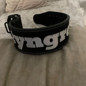 Svart träningsbälte från Tyngre - Säljer ett svart träningsbälte från Tyngre med bred passform. Bältet har stora vita bokstäver på framsidan och flera hål för justering. Perfekt för gymmet när du vill ha extra stöd vid tunga lyft. Det är oanvänt och nypris är 699kr och i storlek XS. Den är tyvärr för liten för mig och där av så säljer jag den. 100% läder (mocka) med snabbspänne.