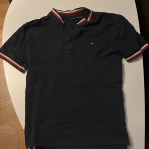Mörkblå t-shirt från Tommy Hilfiger - Mörkblå t-shirt från Tommy Hilfiger med korta ärmar och klassisk ribbad krage. Röd, vit och blå randig detalj på krage och ärmslut. Liten broderad logga på bröstet. Tillverkad i mjuk bomull för en skön känsla.