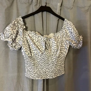 Gråvit leopardmönstrad croppad blus - Säljer en croppad blus i grått och vitt med leopardmönster. Toppen har puffiga korta ärmar, smockad midja och en söt knytning framtill. Perfekt för dig som gillar trendiga detaljer och offshoulder-stil.
