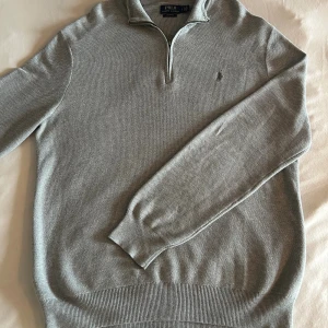 Grå half zip tröja från Polo Ralph Lauren - Snygg grå långärmad tröja från Polo Ralph Lauren med half zip och klassisk krage. Tillverkad i mjuk pima bomull med diskret logga broderad på bröstet. Perfekt för dig som gillar stilrena och bekväma plagg med premiumkänsla.