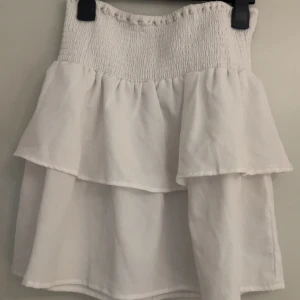 Vit volangkjol från Seaside - Supersöt vit kjol från Seaside med smockad midja och dubbla volanger. perfekt för varma dagar. Enkel att matcha med crop tops eller stickade tröjor för en chill look.