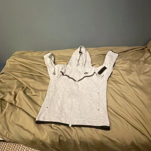 Ljusgrå Nike hoodie med dragkedja - Nike hoodie i ljusgrått med hel dragkedja framtill och huva. Tröjan har svarta detaljer längs dragkedjan och ärmslut, samt en diskret Nike-logga på bröstet. Långärmad och med praktiska fickor på sidorna. Perfekt för chill eller träning.