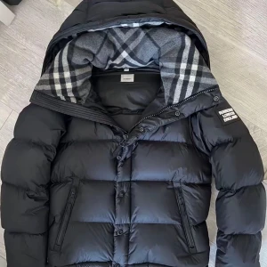 Svart pufferjacka från Burberry - Säljer en svart pufferjacka från Burberry med stor huva och rutig grå/vit insida. Jackan har dragkedja, knappar och en tydlig Burberry London England patch på ärmen. Perfekt för kalla dagar med sin tjocka och fluffiga design.