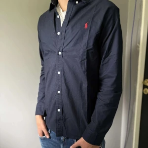 Mörkblå skjorta från Ralph Lauren - Snygg mörkblå skjorta från Ralph Lauren med klassisk krage och vita knappar. Skjortan har lång ärm och ikonisk röd logga broderad på bröstet. Tillverkad i mjuk bomull som känns skön mot huden. Perfekt för dig som gillar stilrena och tidlösa plagg. Hör av dig vid frågor och funderingar😃