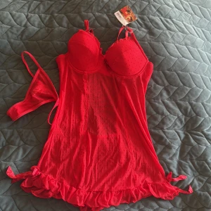 Röd prickig babydoll från Baci Lingerie - Säljer en röd babydoll från Baci Lingerie med prickigt mönster, vadderade kupor och justerbara axelband. Volangkant nertill och små rosetter på sidorna. Matchande stringtrosa ingår. Perfekt för dig som vill ha något extra snyggt och lekfullt.