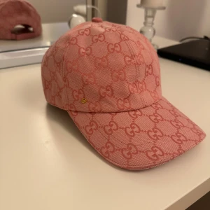 Rosa keps med GG-mönster - Säljer en snygg rosa keps med klassiskt GG-mönster över hela kepsen. Justerbar rem baktill i rosa skinn med guldfärgad metallring. Kepsen har böjd skärm och är tillverkad i bomullstyg med exklusiv känsla. Perfekt för dig som gillar lyxiga accessoarer.