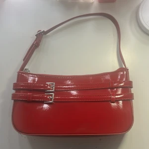 Röd axelväska med spänndetaljer - Snygg röd axelväska i glansigt syntetmaterial med två dekorativa spännen framtill. Väskan har justerbar axelrem och stängs med dragkedja. Liten storlek för det viktigaste och en stilren look som sticker ut.