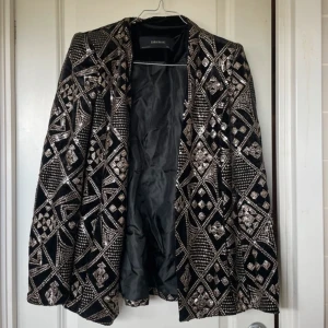Paljettkavaj från Zara Basic, svart/silver - Snygg svart kavaj från Zara Basic med glittriga silverpaljetter i geometriskt mönster. Kavajen har klassisk passform, helfodrad insida och långa ärmar. Perfekt för dig som vill sticka ut med en statement-jacka.