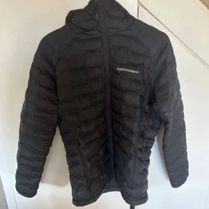 Svart dunjacka Peak Performance - Svart dunjacka från Peak Performance med huva och dragkedja framtill. Jackan är quiltad och har två fickor med dragkedja. Perfekt för kyliga dagar och har en stilren look med diskret logga på bröstet.