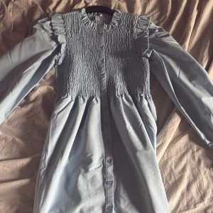 Ljusblå volangklänning från ONLY - Supersöt ljusblå klänning från ONLY med långa ärmar, volangdetaljer vid axlarna och smockad överdel. Klänningen har knappar framtill och är i ett mjukt, lätt material som känns skönt mot huden. Perfekt för dig som gillar en feminin och trendig stil.