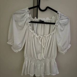 Vit blus med puffärm och volang - Supersöt vit blus från gina tricit med puffiga korta ärmar, volangkant och smockad midja. Toppen har en söt knytning framtill och rynkade detaljer längs halsringningen. 