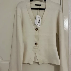 Beige/ crem vit kofta från Gina Tricot - Snygg beige kofta från Gina Tricot med v-ringning och två stora knappar framtill. Koftan har lång ärm och en rak, något kortare passform. Perfekt att slänga över en topp för en chill look. Slutsåld!