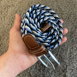 Randigt flätat bälte i blått och vitt - Säljer ett snyggt flätat bälte med randigt mönster i blått, vitt och beige. Bältet har detaljer i brunt skinn och ett silvrigt spänne. Perfekt accessoar för att lyfta din outfit med lite färg och stil.