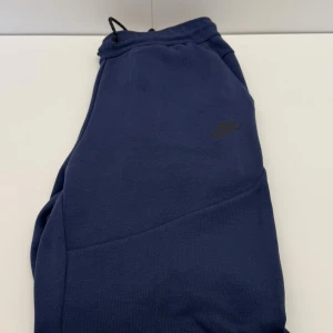 Mörkblå mjukisbyxor från Nike tech - L - Mörkblå mjukisbyxor från Nike med diskret logga på låret. Byxorna har snörning i midjan och dragkedjeförsedda fickor. Tillverkade i mjukt bomullsmaterial för maximal komfort och avslappnad stil.