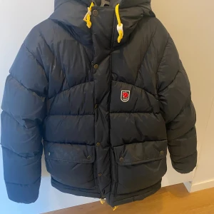 Svart dunjacka från Fjällräven - Svart dunjacka från Fjällräven med gul snörning i huvan och klassisk logga på bröstet. Jackan har stora fickor framtill, dragkedja och tryckknappar. Puffig modell med huva och quiltad design, perfekt för kalla dagar. Pris kan diskuteras 