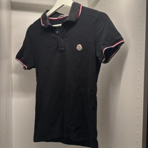 Svart Moncler piké med rödvit kant - Svart pikétröja från Moncler med korta ärmar och klassisk krage. Tröjan har snygga rödvita detaljer på krage och ärmslut samt Moncler-logga på bröstet. Tillverkad i bomull för en skön och stilren känsla.