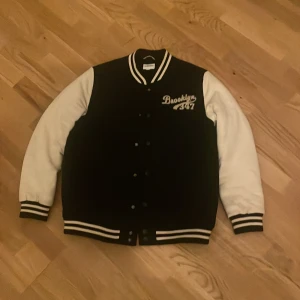 Svart och vit bomberjacka Brooklyn - Snygg svart och vit bomberjacka med broderad Brooklyn 347-logga på bröstet. Jackan har kontrasterande vita ärmar, ribbade muddar och krage med svarta och vita ränder samt knappar framtill. Perfekt för en sportig och trendig look.