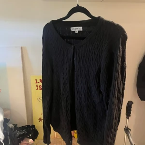 Svart kabelstickad kofta Basic You XL - Svart kabelstickad kofta från Basic You i ekologisk bomull. Koftan har rund hals, knäppning framtill med svarta knappar och långa ärmar. Perfekt för lager-på-lager och passar till allt. Snygg och enkel att styla till olika looks.