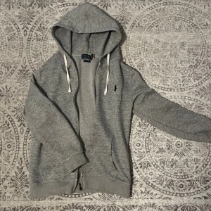 Grå hoodie från Polo Ralph Lauren - Snygg grå hoodie från Polo Ralph Lauren med svart logga på bröstet. Tröjan har huva med vita snören, dragkedja framtill och två fickor. Tillverkad i mjuk bomullsmix och har långärmad passform. Perfekt för chill dagar och streetwear. Storlek s men sitter XS