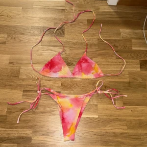 Färgglad bikini med knytband - Aldrig använt Superfin bikini i rosa, gul och orange med tie-dye känsla. Triangeltopp med smala knytband i nacken och ryggen, samt matchande trosa med knyt i sidorna. Perfekt för stranden eller poolen, riktigt somrig vibe!