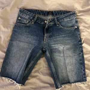 Blå jeansshorts från NLY Jeans - Säljer ett par blå denimshorts från NLY Jeans med låg midja och fransiga benslut. Klassisk femficksmodell med knapp och dragkedja framtill. Materialet är 100% bomull och shortsen har en loose passform. 