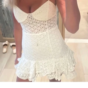 Vit kort spetsklänning med volang - Supersöt vit kort klänning med tunna axelband och v-ringad byst. Klänningen har broderad spets med hålmönster och volangkant nedtill som ger en extra flirty vibe. Perfekt för sommarens alla tillfällen, och den är lätt och luftig i materialet.