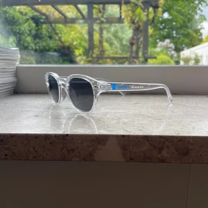 Djurgår'n transparenta solglasögon (Limited edition) - Snygga solglasögon med transparent båge och svarta runda linser. På skalmen finns en blå Djurgår'n-logga som ger extra stilpoäng. Perfekt accessoar för dig som vill sticka ut och visa klubbkänsla. Lätta och bekväma att bära under soliga dagar.