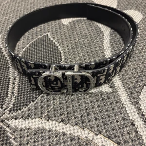 Svart och vit Dior bälte med CD-spänne - Säljer ett snyggt Dior bälte med klassiskt CD-spänne i silver. Bältet har ett grafiskt mönster i svart och vitt med Dior-logga över hela bandet. Insidan är i svart skinn och bältet är stilrent och lyxigt, perfekt för att lyfta din outfit.