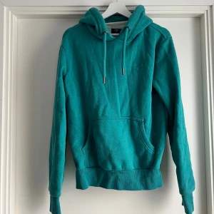 Turkos hoodie från Superdry - Snygg turkos hoodie från Superdry med huva och snörning. Tröjan har en stor magficka och ribbade muddar vid ärmslut och nederkant. Perfekt för chill dagar och har en mjuk insida i fleece som gör den extra skön.