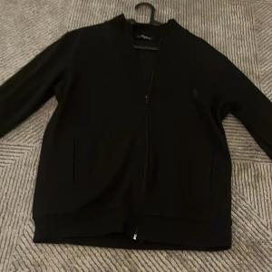 Svart bomberjacka med dragkedja - Svart bomberjacka från Ralph Lauren med stilren design och diskret krage. Jackan har hel dragkedja framtill och två sidofickor. Materialet är mjukt och känns skönt mot huden, perfekt för dig som gillar enkel och clean look.  Har katt hemma så därför syns det lite päls men Hoodien är ny