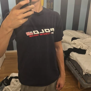 Mörkblå Polo Ralph Lauren t-shirt - Snygg mörkblå t-shirt från Polo Ralph Lauren med broderad text 'POLO US CREW GAME' och amerikansk flagga på bröstet. Klassisk rund hals och korta ärmar. Materialet är mjuk bomull, perfekt för en chill och sportig look. Släpptes limiterad. Den är lite stor i storleken passar stor M till L.