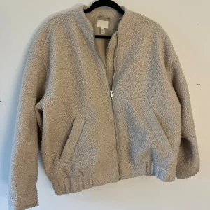 Beige teddyjacka från H&M - Mysig teddyjacka från H&M i beige färg med dragkedja framtill och två snedställda fickor. Jackan har en rundad krage och är tillverkad i fluffigt syntetmaterial som ger en chill vibe. Perfekt för att hålla sig varm under kyliga dagar.