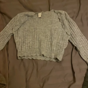 Grå croppad stickad tröja - Supermysig grå croppad stickad tröja med långärmad design och ribbade detaljer. Tröjan har vågiga kanter vid halsringning, ärmslut och nederkant som ger en extra touch. Perfekt för dig som gillar en chill och trendig look.