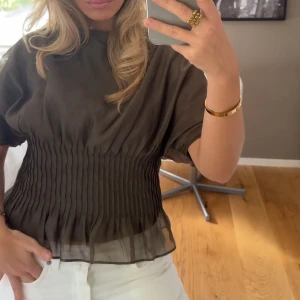 Plisserad grön blus med kort ärm - Snygg grön blus med plisserad nederdel och transparent material längst ner. Blusen har korta ärmar och en lös passform upptill, vilket ger en cool och trendig look. Perfekt att styla med jeans eller kjol. Helt ny med prislapp