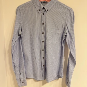 Randig blå skjorta från Terranova - Snygg skjorta från Terranova i storlek M med tunna blå och vita ränder. Klassisk krage, bröstficka och bruna knappar framtill. Långärmad modell i mjuk bomull som känns chill och enkel att matcha. Perfekt för dig som gillar stilrena detaljer och relaxed vibe.